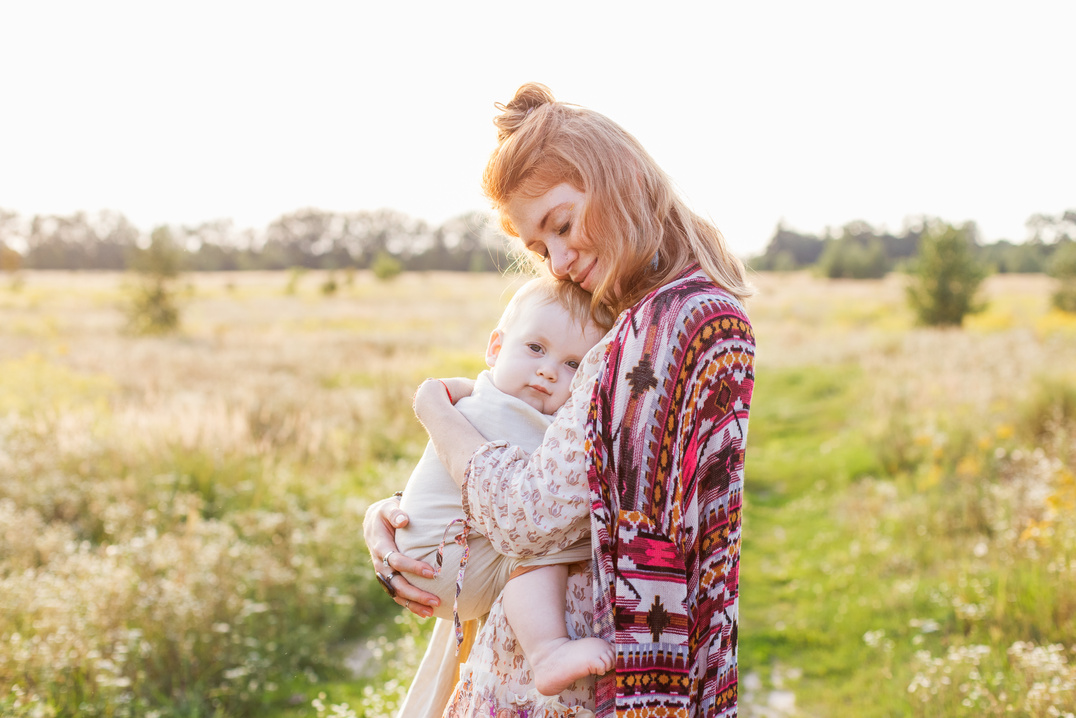 Little baby boy and his mother walking in the fields during summer day. Mother is holding and tickling her baby, babywearing in sling. Natural parenting concept